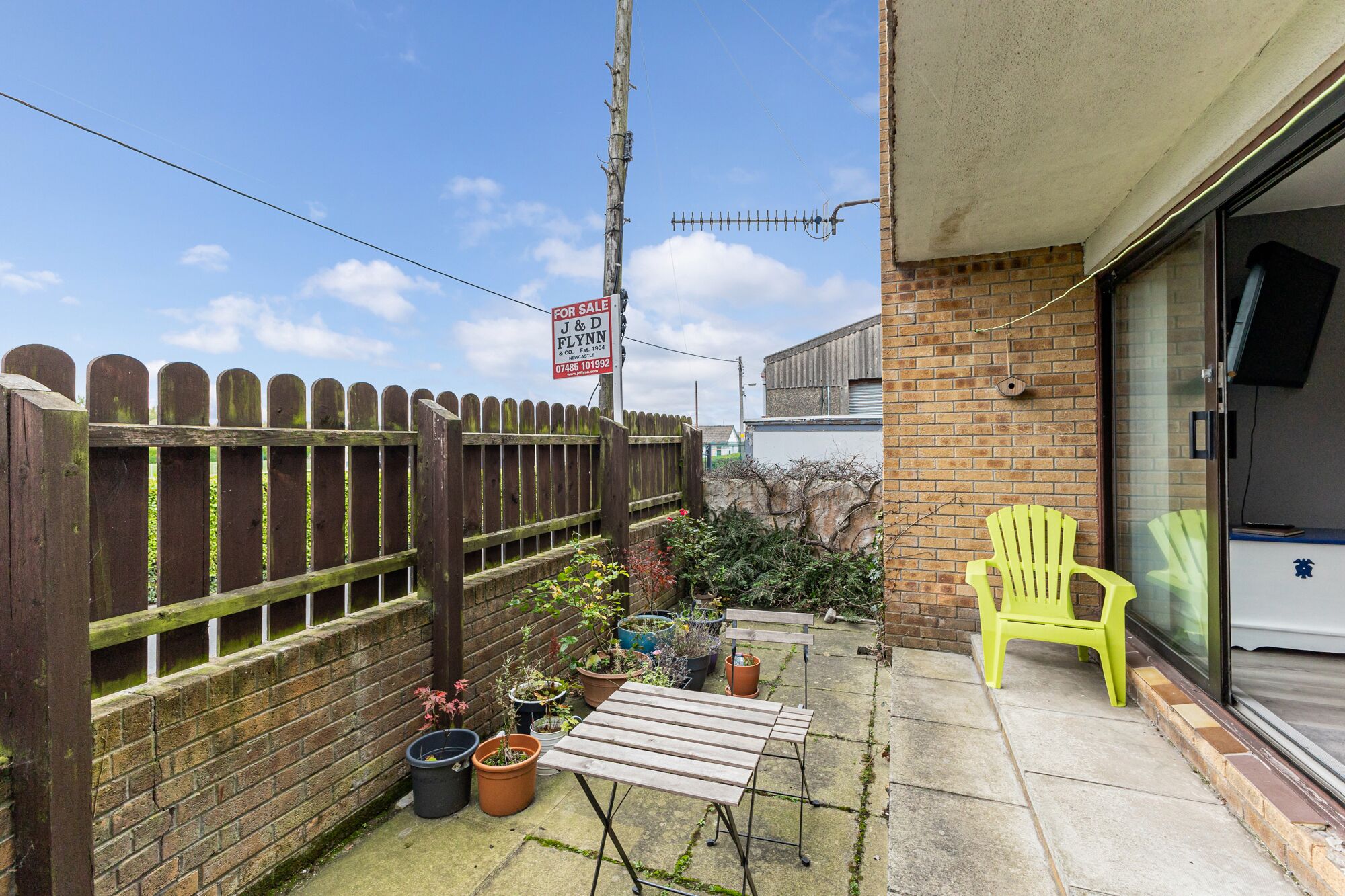 A small outdoor patio area with a wooden table and chairs, potted plants, and a yellow chair.