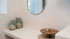 Une salle de bain avec un banc blanc, deux vases verts, un miroir rond et une table en bois sur laquelle se trouve une montre.