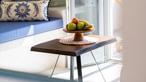 Une table à manger agrémentée d’un centre de table composé de fruits, dans une pièce lumineuse où se trouvent un canapé bleu et des coussins à motifs.