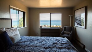Een slaapkamer met een bed, een stoel, een bureau en een groot raam met uitzicht op de oceaan.