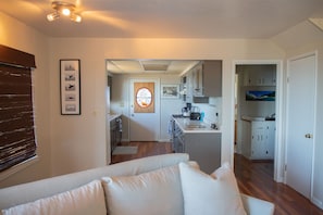 A modern living room with a white sofa, a kitchen with stainless steel appliances, and a dining area with a view of the outdoors. - The Blue Pelican (Whitethorn)