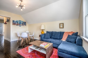A living room with a blue sofa, a wooden coffee table, a red patterned rug, and a dining area with white chairs and a small table. - Modern 3-Bed Stay w/ Patio, Firepit, & Garage Parking Spot (Chicago)