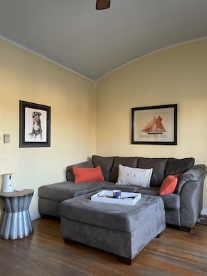 Una sala de estar con un sofá seccional gris, un otoman gris y una pequeña mesa auxiliar con un jarrón. Hay dos cuadros en la pared.