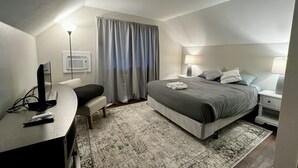 Une chambre à coucher comprenant un lit, des tables de chevet, un bureau, une chaise, une lampe et une fenêtre avec des rideaux.