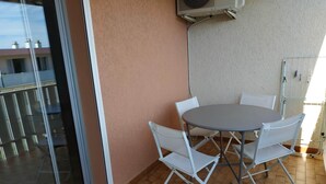 Un balcón con puerta de cristal, una mesa redonda con cuatro sillas blancas y un equipo de aire acondicionado en la pared.