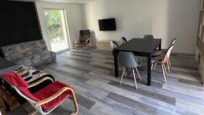 A spacious living area with a large sofa, a dining table with chairs, a television mounted on the wall, and a window providing natural light.