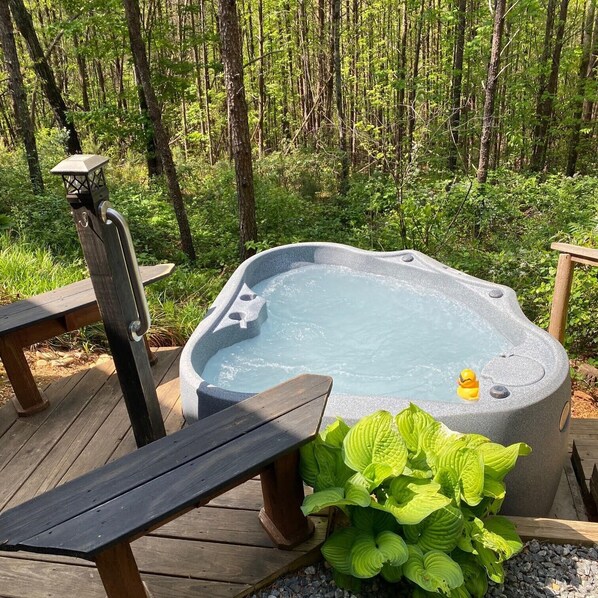 A hot tub in a forested area with wooden deck seating and lush greenery. - Serenity Ridge Cabin F: Couple's Vacay + Hot Tub (Mill Spring)