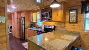 Interior de una cabaña de madera con cocina que incluye encimeras de granito, un refrigerador, un microondas y una estufa. Hay una barra con taburetes y un ventilador de techo.
