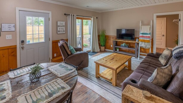 A cozy living room with a brown leather sofa, a wooden coffee table, and a TV on a wooden stand. There is a dining table with placemats and a potted plant. - Hill Hideaway Unit 2 - Pet-Friendly Lake Retreat (Minocqua)