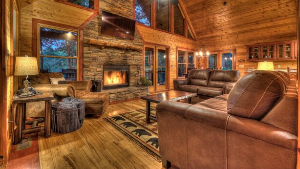 A cozy log cabin interior with a stone fireplace, leather furniture, and large windows offering views of the forest. - Aska Retreat - Pet Friendly | Hot Tub | Foosball | Arcade Games (Blue Ridge)