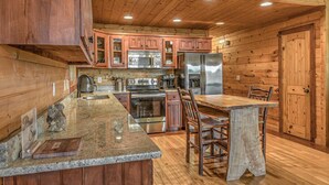 A kitchen with wooden cabinets, stainless steel appliances, granite countertops, and a rustic dining table. - Aska Retreat - Pet Friendly | Hot Tub | Foosball | Arcade Games (Blue Ridge)