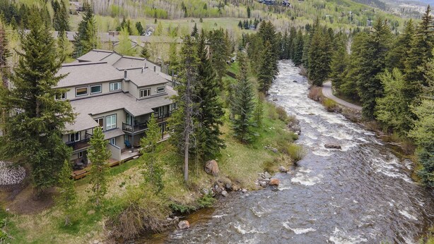 A multi-story house with a river flowing nearby, surrounded by dense forest and hills.