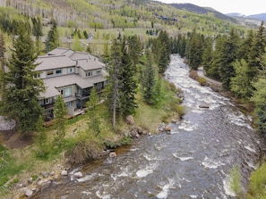 A multi-story house with a river flowing nearby, surrounded by dense forest and hills. - Coldstream Townhome 19 (Vail)