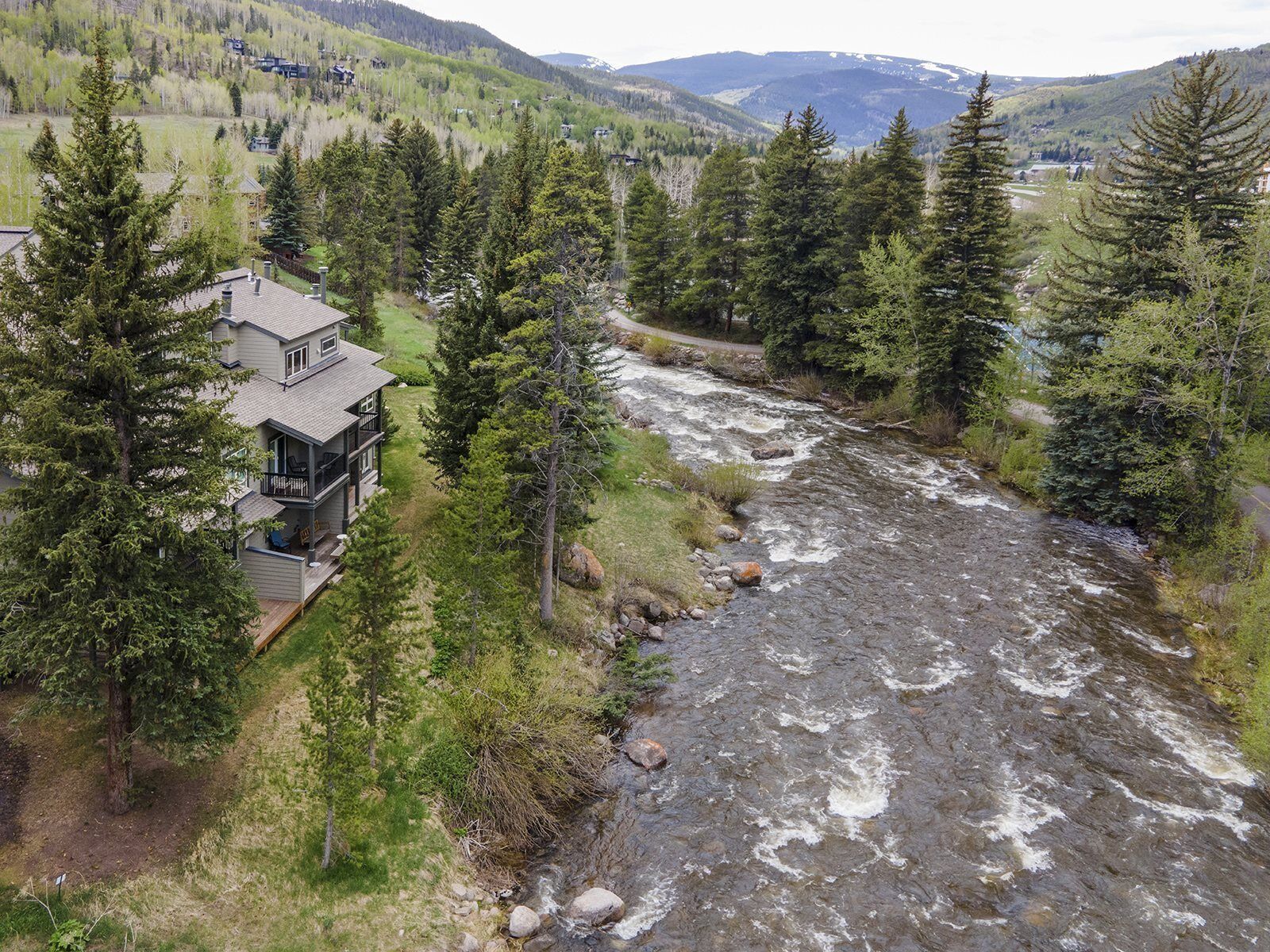 A two-story house with a balcony overlooking a river, surrounded by dense forest and mountains in the background.