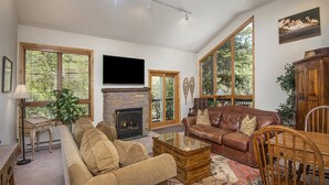 A spacious living room with a stone fireplace, leather sofa, and large windows offering views of trees.