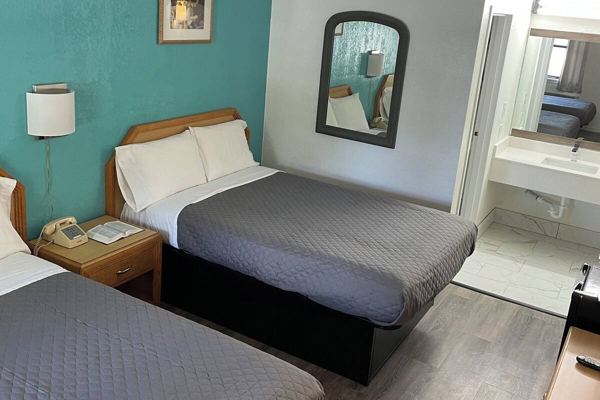 A hotel room with two beds, a bedside table with a lamp, a mirror, and a small desk with a chair.