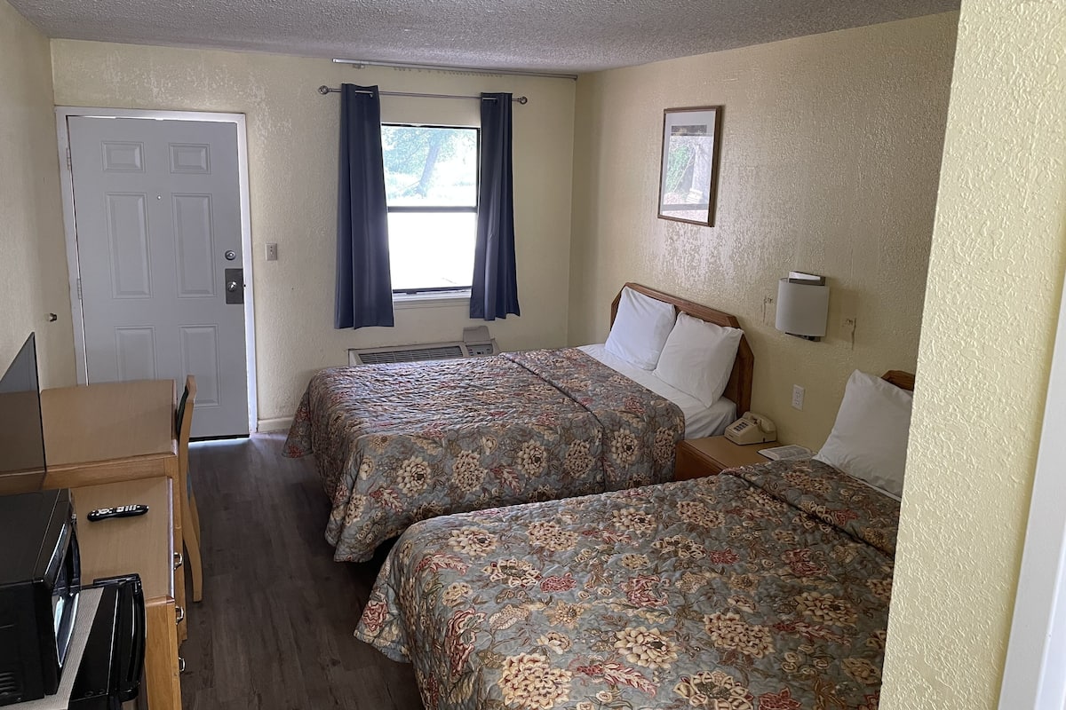 A hotel room with two beds, a desk, a chair, a window with curtains, a door, and a framed picture on the wall.