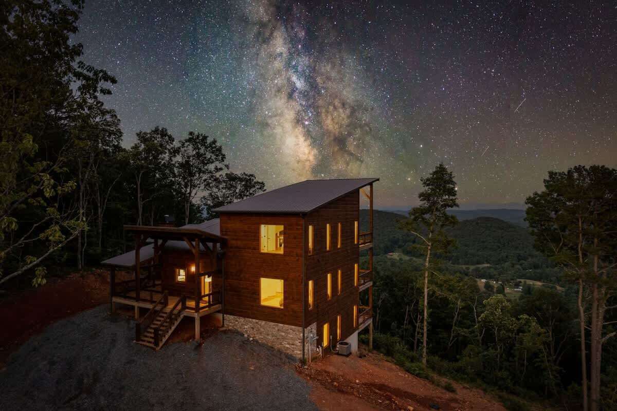 A wooden cabin with multiple windows and a staircase, surrounded by trees and a starry night sky.