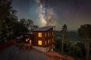 Une cabane en bois dotée de nombreuses fenêtres et d’un escalier, entourée d’arbres et surplombant un ciel étoilé.