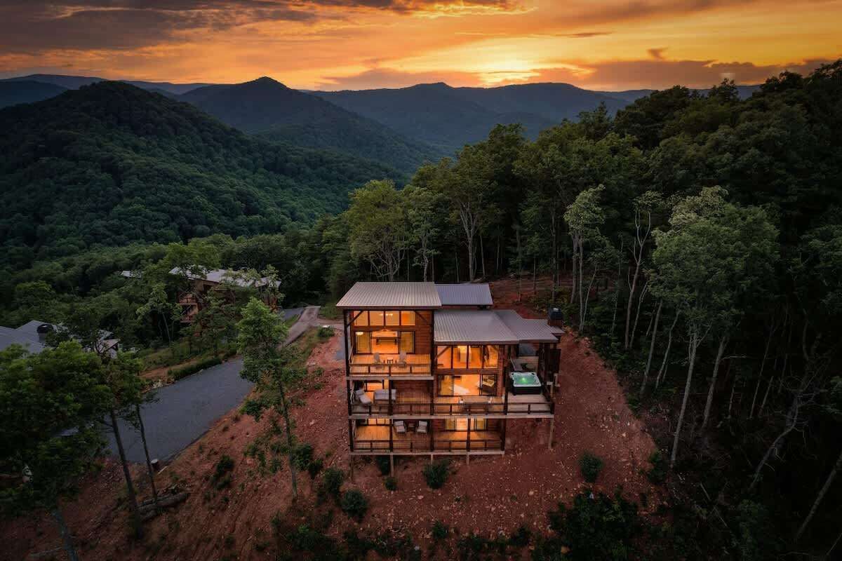 A two-story wooden cabin with a balcony, surrounded by forested hills.