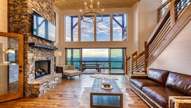 A spacious living room with a stone fireplace, leather sofa, and large windows offering a view of the outdoors. - Big Sky 2 - Hot tub & game room (Epworth)