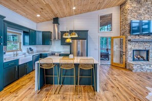 A modern kitchen with a central island, bar stools, and a stone fireplace. - 6 Bedroom Mountain Cabin w/ Hot Tub & Game Room (Epworth)