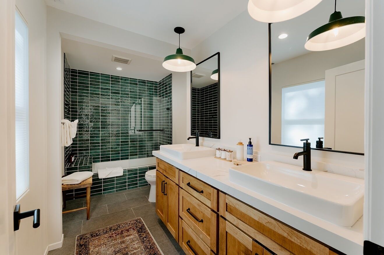 A modern bathroom with a double sink vanity, green-tiled shower, and pendant lighting.