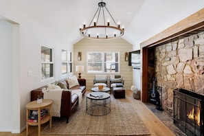 A cozy living room with a stone fireplace, a brown leather sofa, a round coffee table, and a chandelier.