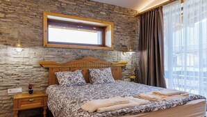 A bedroom with a stone wall, a wooden headboard, a bed with floral bedding, a nightstand, and a window with curtains. - Villa Raphael near Pirin Golf Resort (Razlog)