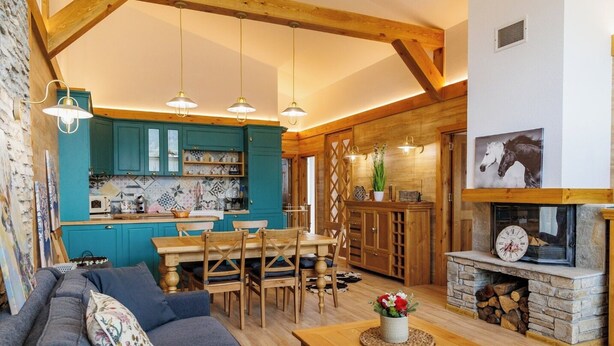 A cozy living room with a sofa, wooden dining table, and chairs, a kitchen with teal cabinets, and a fireplace with a clock. - Villa Raphael near Pirin Golf Resort (Razlog)