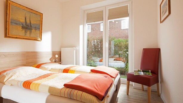 A hotel room with two beds, a red chair, and a large window with a view of greenery. - Ferienhaus Mien Huske (Norderney)