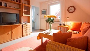 A cozy living room with a television, bookshelf, and a window with curtains. - Ferienhaus Mien Huske (Norderney)