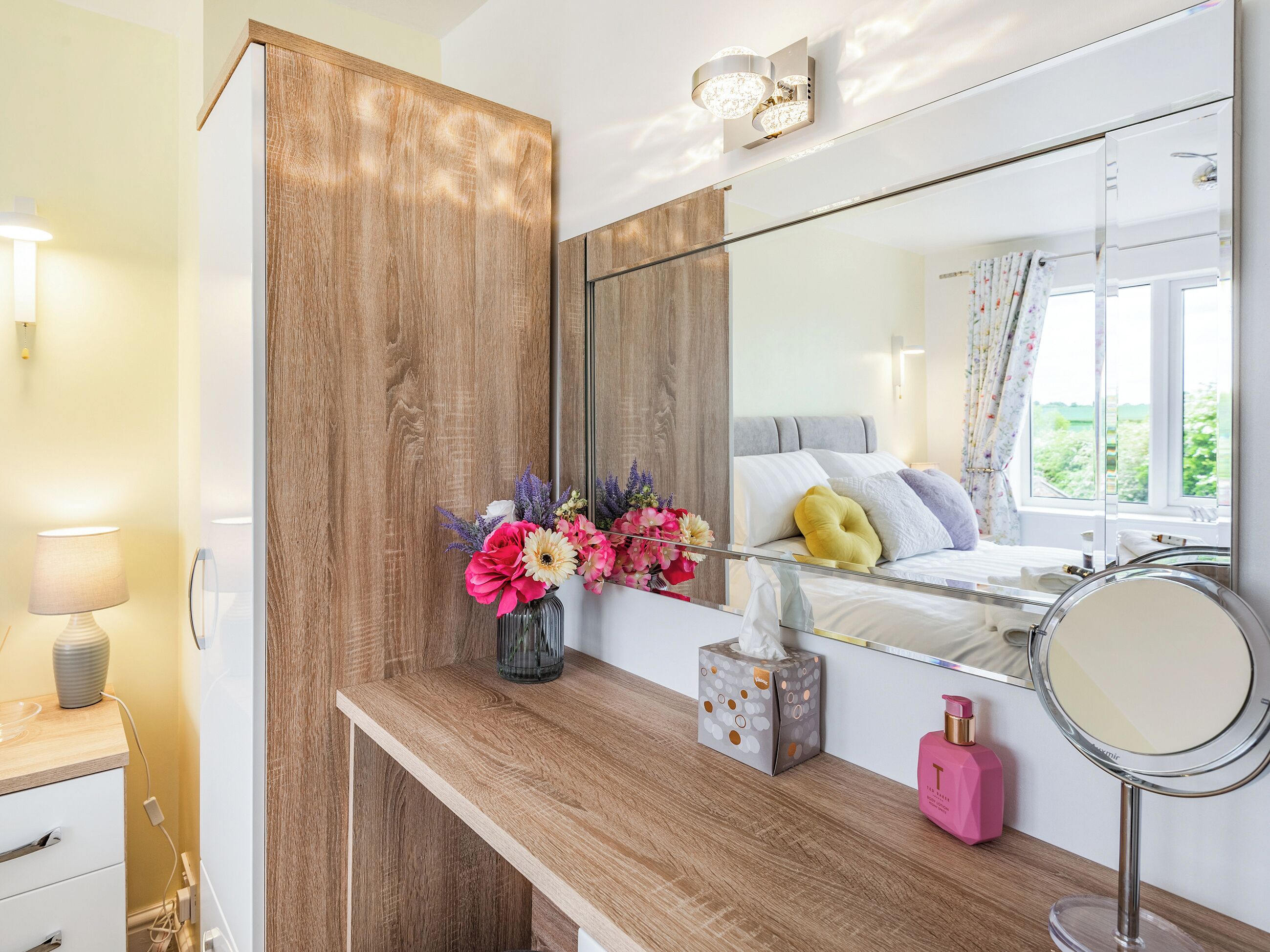 A bedroom with a wooden vanity, a mirror, a bedside table with a tissue box, a pink bottle, and a window with curtains.