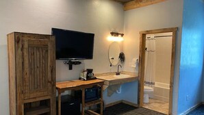 Un baño pequeño con tocador de madera, televisor montado, lavamanos con espejo y zona de ducha.