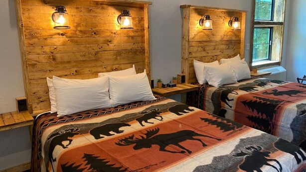 Dos camas con cabeceros de madera y lámparas, una ventana con vistas a follaje, y una manta estampada en una cama.