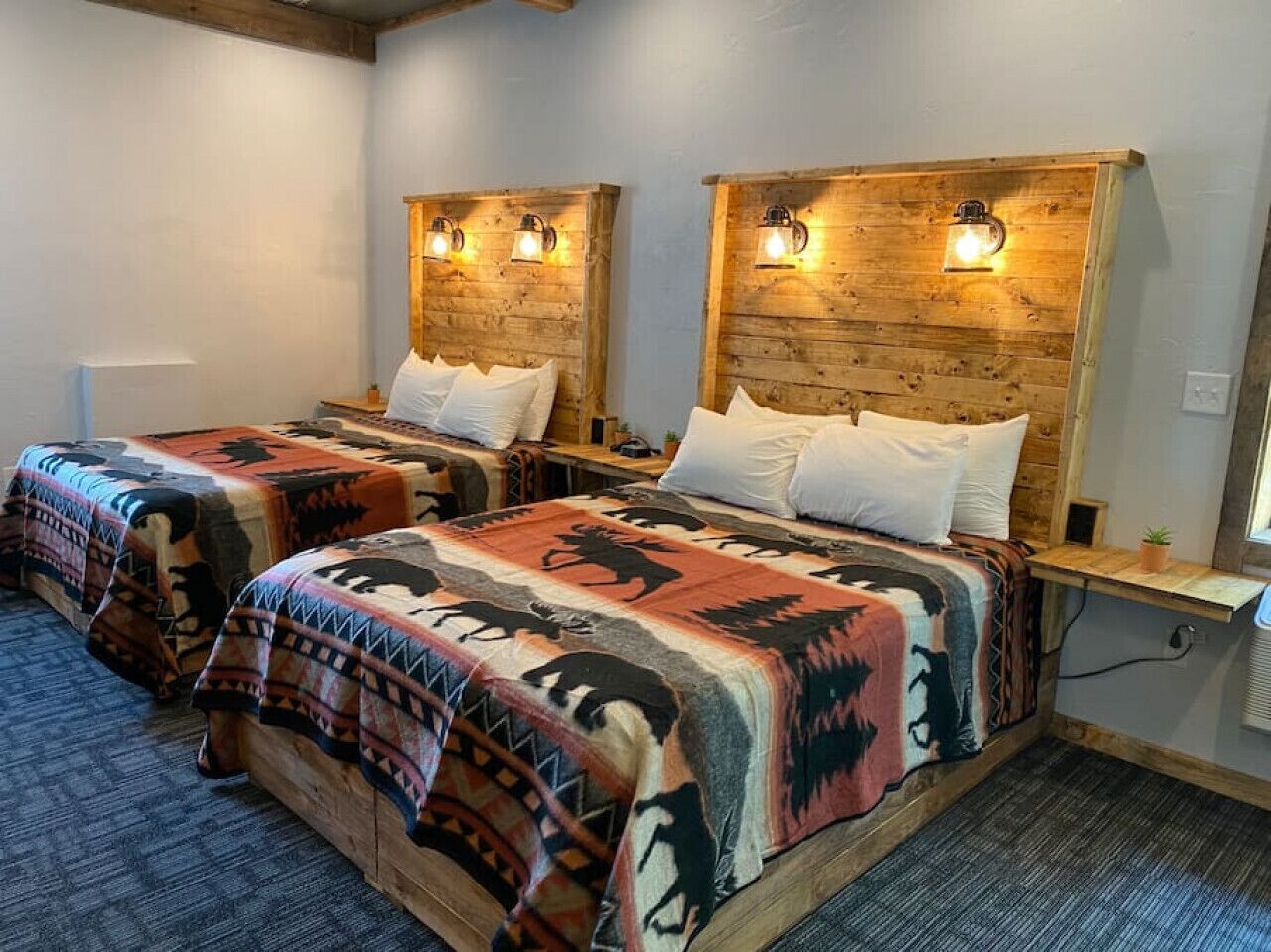 Two twin beds with patterned blankets, wooden headboards with wall-mounted lights, and a small bedside table with a plant.
