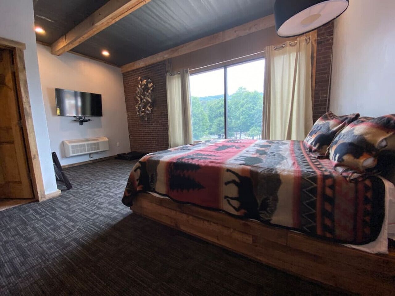 A bedroom with a large bed, a flat-screen TV mounted on the wall, a window with curtains, and a brick wall.