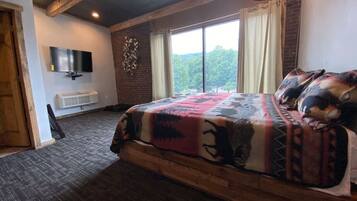 A bedroom with a large bed, a flat-screen TV mounted on the wall, a window with curtains, and a brick wall.