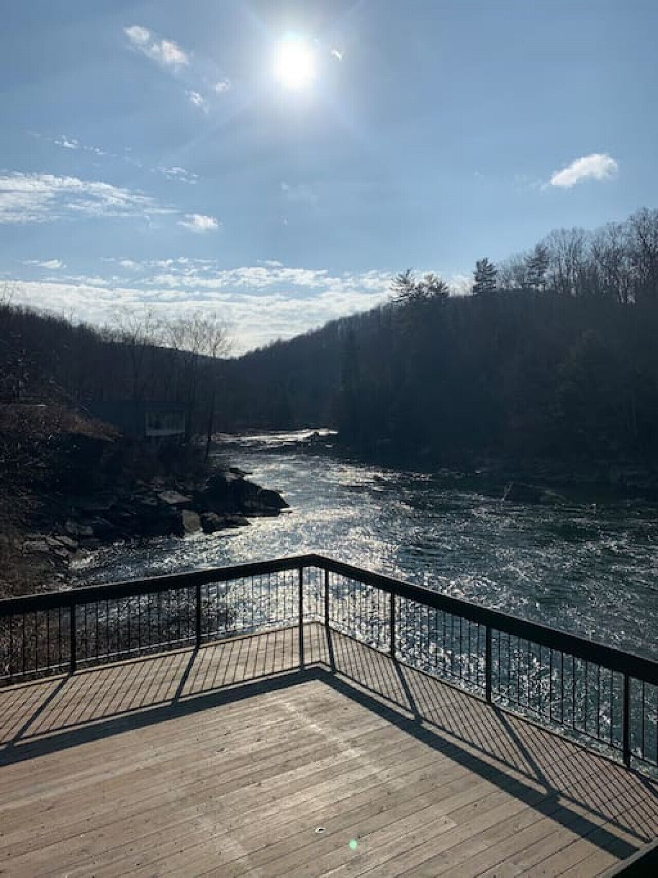 A wooden deck with a metal railing overlooking a river, surrounded by trees and a clear sky.