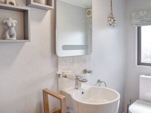 Ein Badezimmer mit einem weißen Waschbecken, Spiegel und Toilette. Regale mit Dekorationselementen und ein Fenster mit Jalousien.