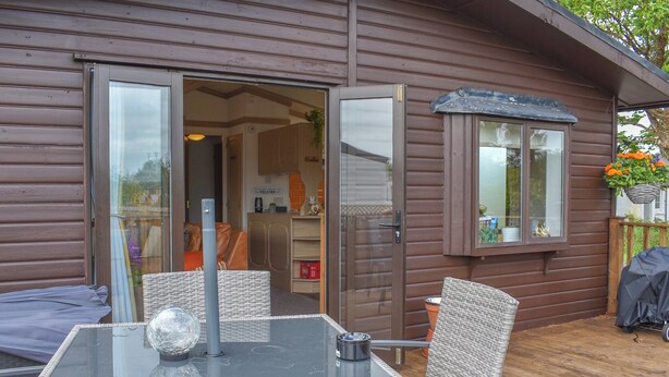 A glass-top table with wicker chairs in a wooden deck area of a cabin. - Y Nyth Yn Bwthyn Coed Gellyg (Dinas Dinlle, near Caernarfon)