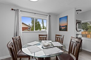 A dining area with a glass table surrounded by wooden chairs, a white ceiling, and a window with curtains.