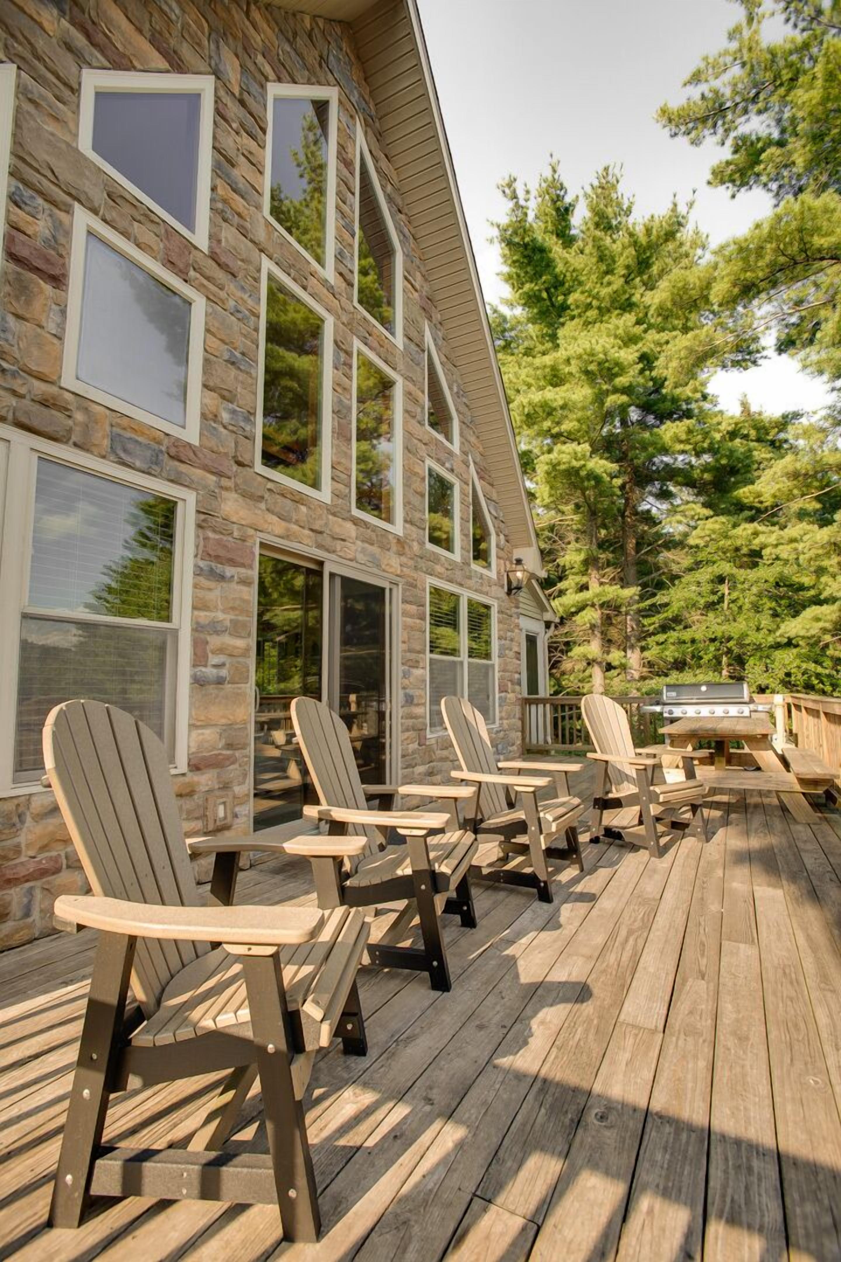 A wooden deck with Adirondack chairs and a grill, overlooking a stone building with large windows.