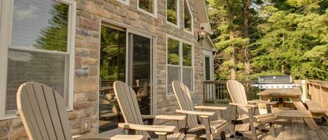 A wooden deck with Adirondack chairs and a grill, overlooking a stone building with large windows.