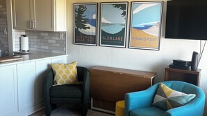 A modern living room with a sofa, ottoman, and a bench. There are framed pictures on the wall, a flat-screen TV, and a kitchen area with cabinets and a countertop.