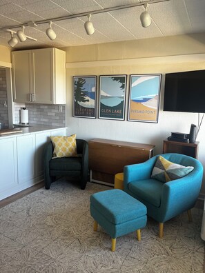 A modern living room with a sofa, ottoman, and a bench. There are framed pictures on the wall, a flat-screen TV, and a kitchen area with cabinets and a countertop.