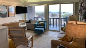 Ein Zimmer mit Schreibtisch, Stuhl, einer Couch und einem großen Fenster mit Blick auf den Strand.