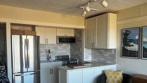 A compact kitchen with white cabinets, a stainless steel refrigerator, a microwave, and a stove. There is a small island with a cutting board, a chair, and a pillow. The floor has a patterned rug, and the ceiling has recessed lighting.