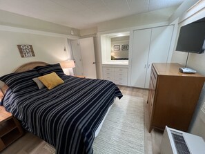 Un dormitorio con una cama, un aparador de madera, un escritorio, una lámpara, un espejo y un televisor sobre un soporte.