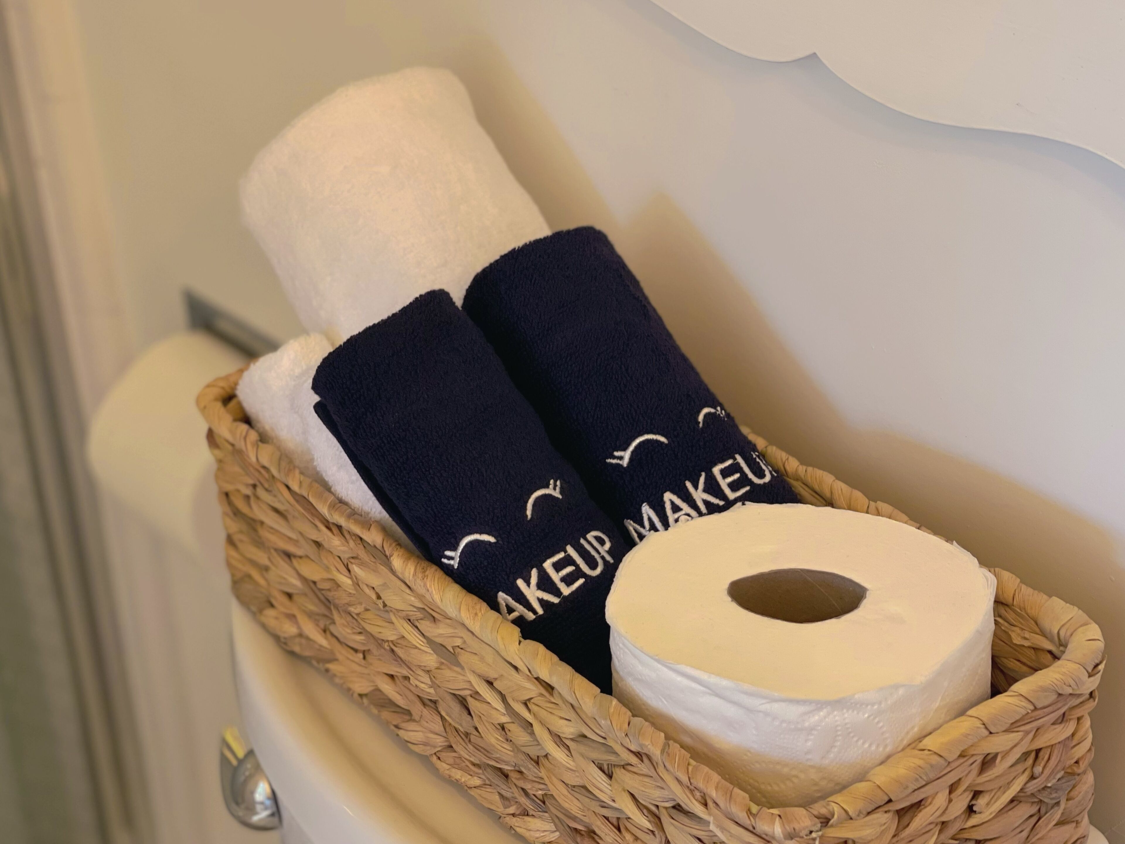 Un panier en osier contenant un rouleau de papier toilette blanc, deux serviettes bleu marine ornées de l’inscription « MAKEUP MAKEUP », et un objet argenté.